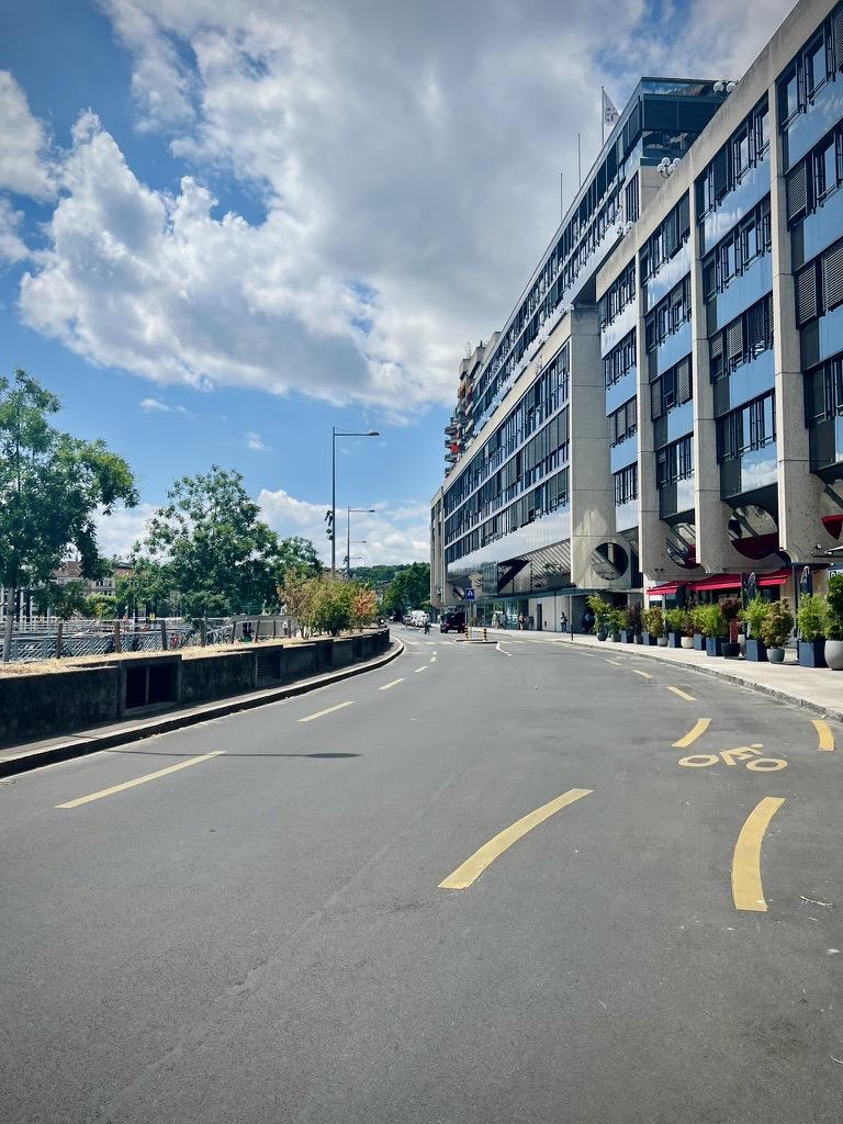 ‎⁨Geneva⁩, ⁨Canton of Geneva⁩, ⁨Switzerland⁩

A wide, curving city road features yellow lane markings and a painted bicycle symbol on the asphalt. On the right, a long modern building with a glass-and-concrete facade is lined with sidewalk planters and shops with red awnings. On the left, a low stone wall separates the road from a line of green trees under a bright blue sky filled with fluffy white clouds.