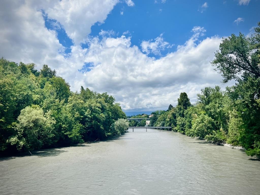 ‎⁨L'Arve⁩, ⁨Geneva⁩, ⁨Canton of Geneva⁩, ⁨Switzerland⁩

A wide, grey-green river flows between dense, leafy green forests that line both banks. In the far distance, a low flat bridge and a higher arch bridge span the water, set against a vibrant blue sky filled with large, billowing white clouds.