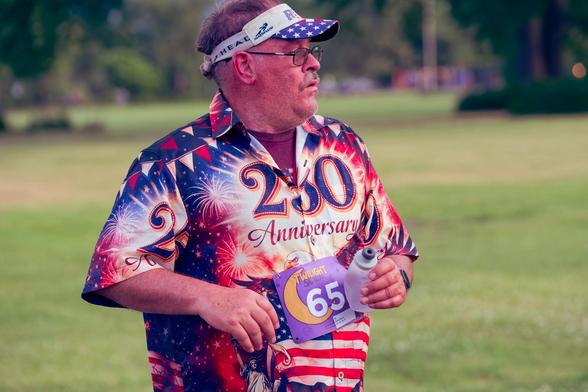Man with partially grey hair wears visor with blue field and with starts and flag-inspired shirt that reads "250- Anniversary" with close croppsed grass and I think the bases of two big willow trees in the background