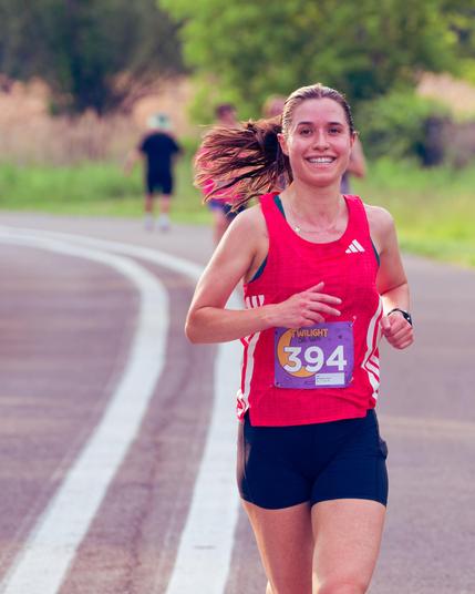 Woman with red adiddas tank top and black shorts has bib number 394, a big smile and hair splayed out to the left with a few runners blurred out behind her
