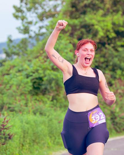 Woman with small black halter top and elastic shorts has an open midriff, reddish hair tied back, airpods, bib number 392, her tounge out and an arm held high with a tatto on her bicep,  a bit of paved trail is in the background in the lower right and a bit of lake, hill and sky in the upper level and the rest is a mass of green vegetation