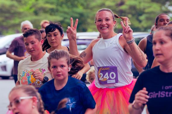 Woman with 240 bib number wears a white shirt and holds up hands in two V's wearing a translucent red-orange skirt over her running gear,  she's surrounded by about 10 other visible runners and some cars can be seen behind them and then some trees.