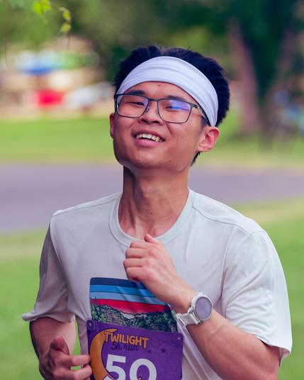 Asian man is smiling  with a big white hairband around his black hair,  his arms are held up in front of him, one with a watch and he's wearing an off-white t-shirt with a graphi mostly covered by his number 50 bib.  There is short cropped grass behind him with a paved path crossing horizontally need the center, above that a big tree to the right and spots of red and blue (playground equipment?) to the left