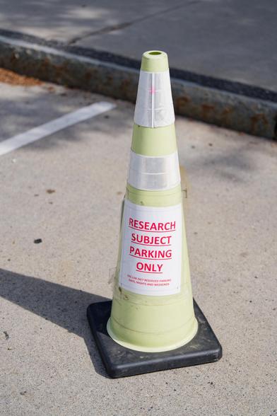 I moved this traffic cone to a good spot on the road by a sidewalk,  it is light yellow, not orange, and has a white sign attached that reads in red underlined text RESEARCH SUBJECT PARKING ONLY