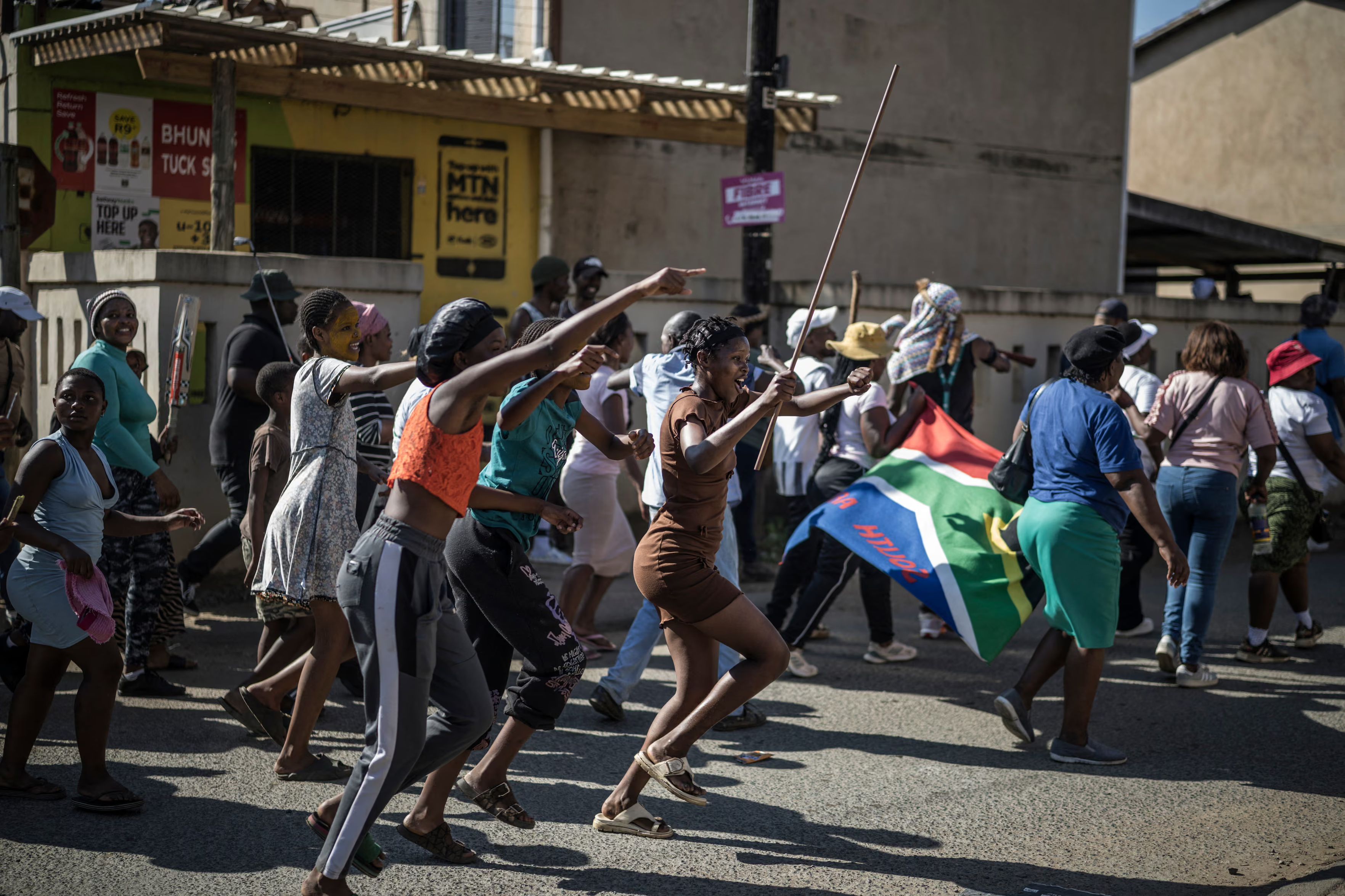 A dynamic, eye-level photograph of a crowd of people marching and running down a paved street during a sunny day. In the foreground, several young women are moving energetically forward; one points ahead with an outstretched arm, while another raised a long thin stick in the air. In the mid-ground, a South African national flag is held low by marchers. The background features single-story storefronts with advertising signs ("BHUNGU TUCK SHOP", "MTN here") and other pedestrians joining or watching the demonstration.