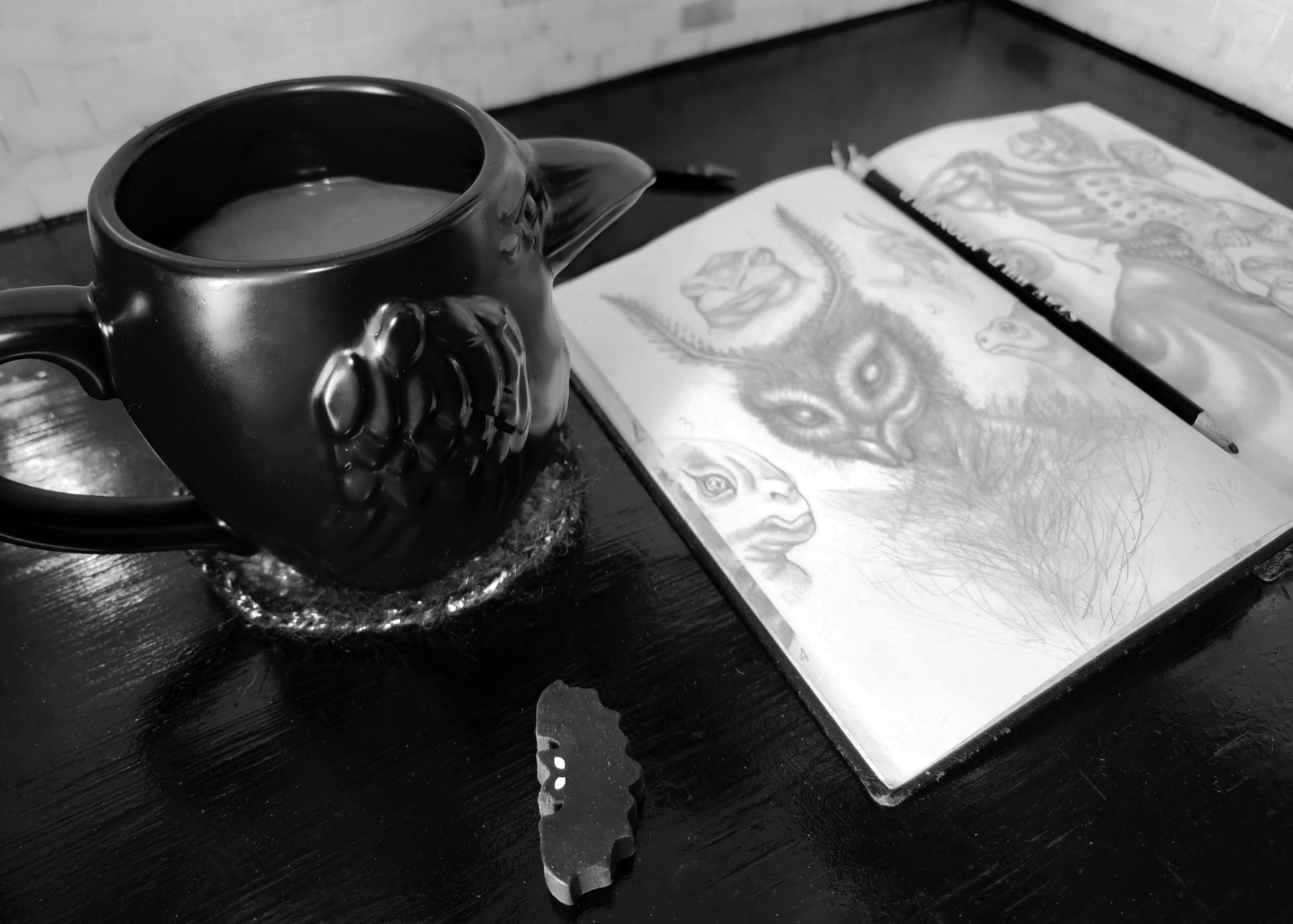 A black and white photo of a ceramic raven mug with a 3D wing and beak design sitting on a dark wooden table next to an open sketchbook with doodles of alien creatures on the pages. A pencil rests on the book, with a bat-shaped eraser resting on the side.