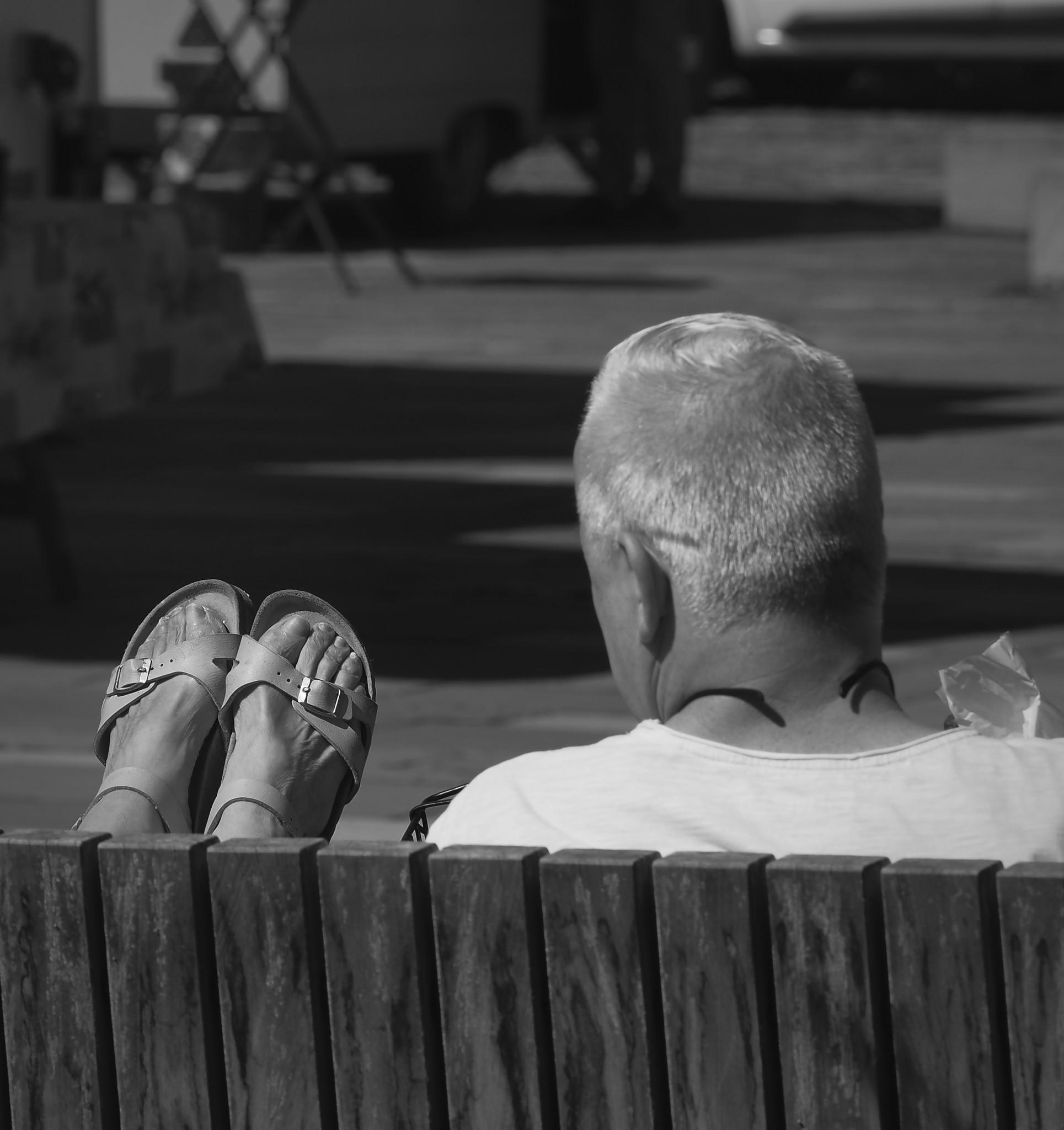 Von hinten aufgenommen: Ein Mann sitzt auf einer Bank und schaut in Richtung der hochgelegten Füße seiner Sitznachbarin.