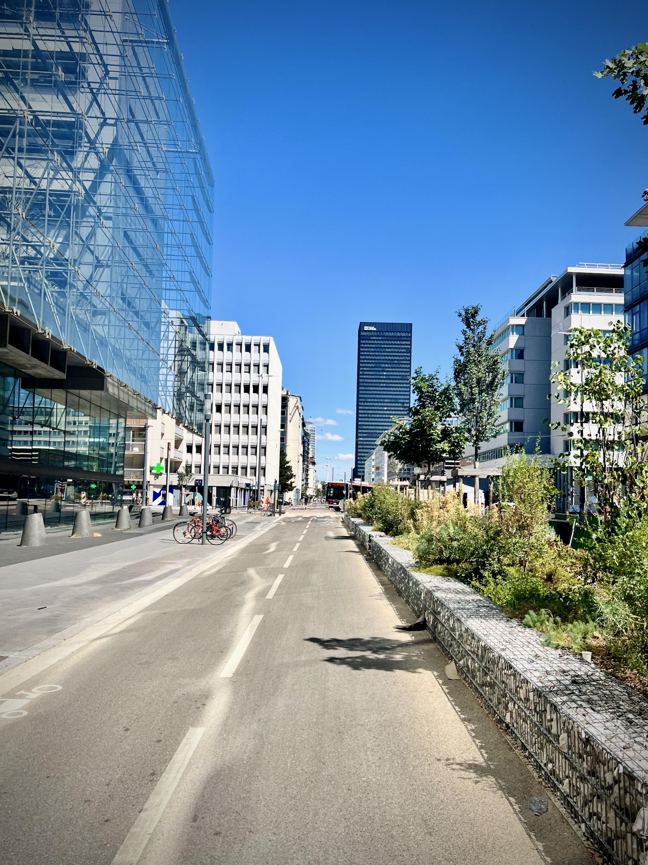 ‎⁨Near to Part-Dieu, Lyon⁩, ⁨Auvergne-Rhône-Alpes⁩, ⁨France⁩

A wide, straight paved bike path runs through a modern city district, bordered by a low stone-filled gabion planter with green shrubs on the right. On the left, a large glass-fronted building stands beside a pedestrian sidewalk, while a tall, dark monolithic skyscraper dominates the far horizon under a clear blue sky.