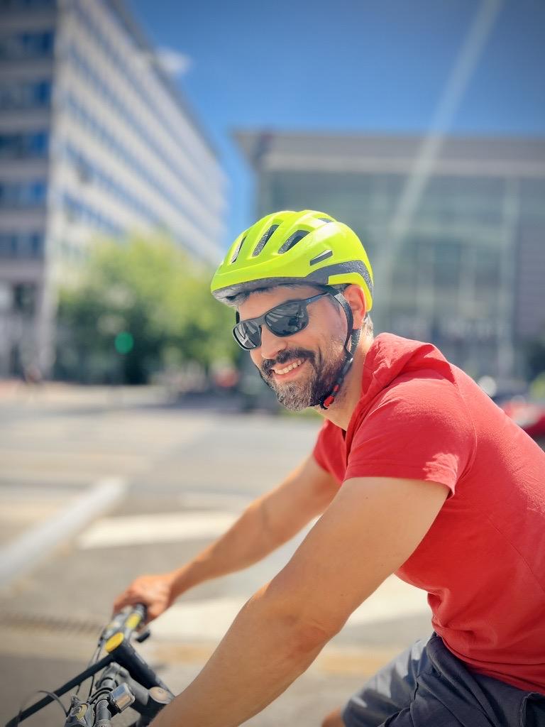 ‎⁨Lyon⁩, ⁨Auvergne-Rhône-Alpes⁩, ⁨France⁩

A close-up portrait of a smiling, bearded man (Pierre) riding a bicycle and looking back over his shoulder toward the camera. He wears a bright neon-yellow helmet, black sunglasses, and a red t-shirt, with a soft-focus city street and modern buildings in the background under bright, sunny daylight.