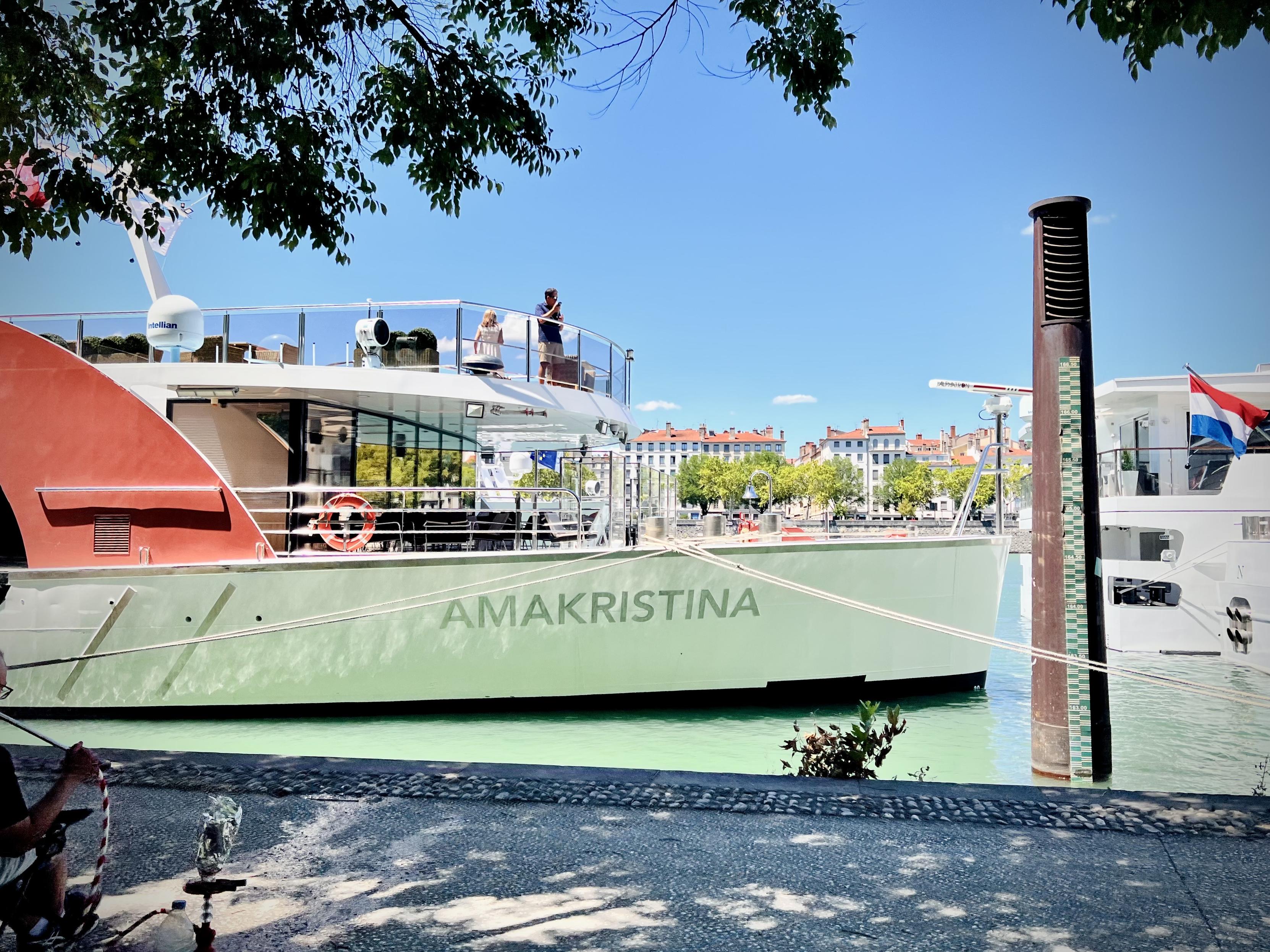 Quays of the Rhône, ‎⁨Lyon⁩, ⁨Auvergne-Rhône-Alpes⁩, ⁨France⁩

A large, light-green river cruise ship named "AMAKRISTINA" is docked along a paved city quay, with a couple of people standing on its upper sun deck. In the background, elegant European buildings line the far riverbank under a clear blue sky, while a tall metal mooring pillar with a water level gauge stands on the right.