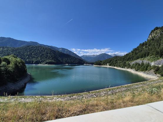 This image captures a breathtaking and expansive mountain lake scene, showcasing the serene beauty of a high-altitude landscape. The foreground features a vibrant, turquoise-colored lake, its calm waters reflecting the clear blue sky above. The lake is nestled between steep, forested slopes, creating a stunning contrast between the water and the lush greenery.
The surrounding mountains are covered in dense forests of evergreen trees, their dark green foliage adding depth and texture to the scen…