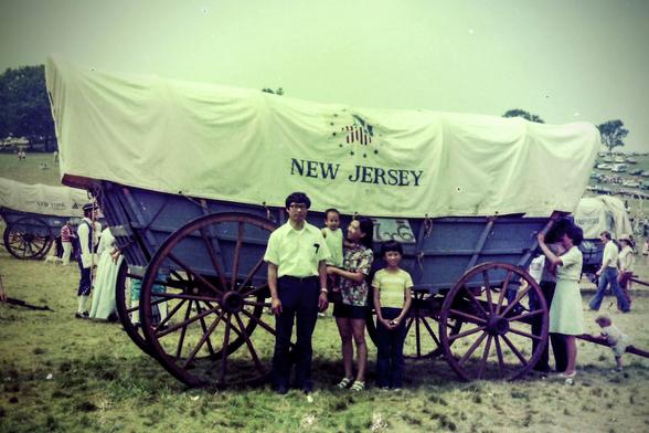 Scan of a photo from 1976, tinted greenish-yellow with age.

In front of a covered wagon with the words "New Jersey" on its canopy, there is a family of four. Dad has glasses, a white shirt with a glasses case in the breast pocket, and dark slacks. Mom is wearing a colorful patterned blouse, a short dark skirt, and white strap sandals. She's holding up a toddler wearing a light-colored onesie. Next to her stands a preteen boy wearing dark pants and a shirt that looks like it has yellow and wh…