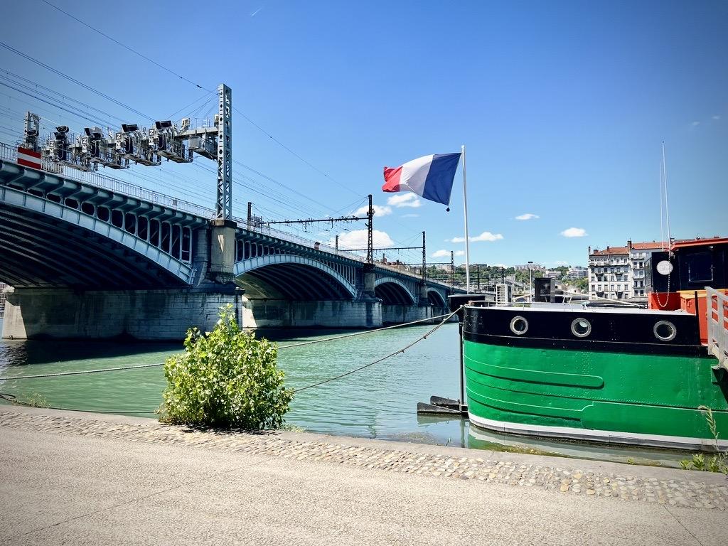 ‎⁨Lyon⁩, ⁨Auvergne-Rhône-Alpes⁩, ⁨France⁩

A French flag waves from a green-hulled barge moored along a paved riverbank. In the background, a large, multi-arched railway bridge with overhead electrical lines spans the river under a clear blue sky on a bright, sunny day.