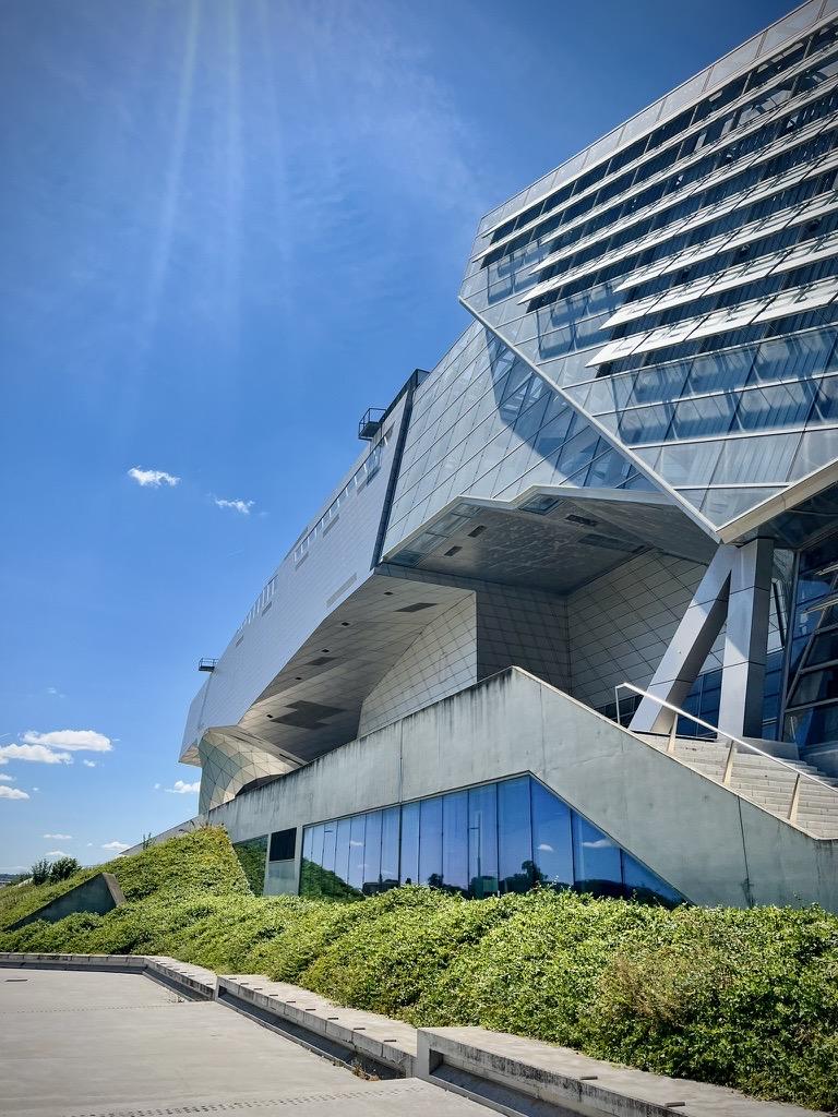 ‎⁨Jardin du Musée des Confluences⁩, ⁨Lyon⁩, ⁨Auvergne-Rhône-Alpes⁩, ⁨France⁩

A low-angle view of an ultra-modern building featuring complex geometric architecture with cantilevered steel and glass facades. In the foreground, concrete walkways, stairs, and low walls are bordered by landscaped green shrubbery under a bright, clear blue sky.