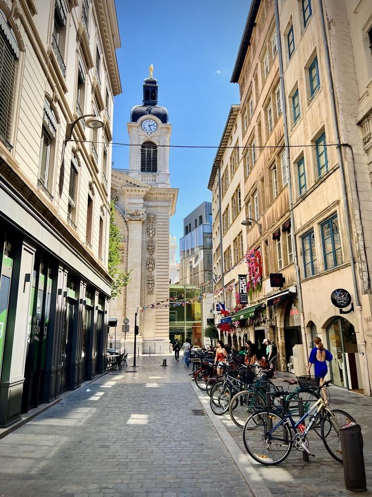 Rue Confort, ‎⁨Lyon⁩, ⁨Auvergne-Rhône-Alpes⁩, ⁨France⁩

A narrow, paved pedestrian street is framed by tall historic buildings, with parked bicycles and outdoor cafe seating along the right side. In the background, a prominent stone clock tower with a blue clock face and a black domed top rises at the end of the street beneath a clear blue sky.