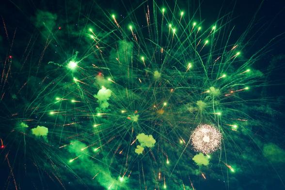 Lots of green burning dots trailing lines of smoke that seems to mostly radiate from a center but to also kinda go out in al directions need the center with bigger diffuse clouds of smoke lit up green by the fireworks and also towards the lower right a white expanding firework looks like a small dandilion puff