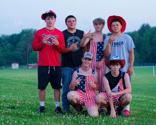 6 young men pose in clover-studded lawn in front of athletic field and dark green hedgerow.  4 in the back,2 in the middle,  and two wear matching flag dungaress.
