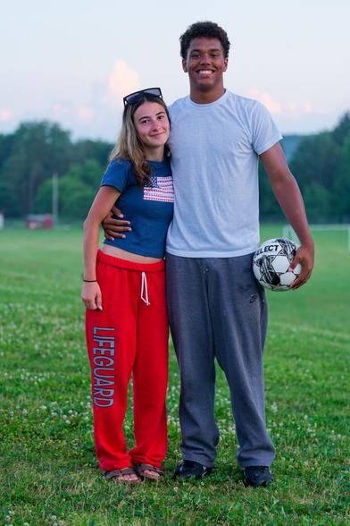 On the left a white woman wearing red pants that say lifeguard and a blue shirt with an american flag and open midriff,  to the right a taller black man with grey pants and with t-shirt holding a soccer ball, both smiling.
