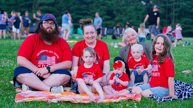 Mutigenerational family on blanket all wearing red shirts with an american flag celebrating our 250th year with more people in the far distance