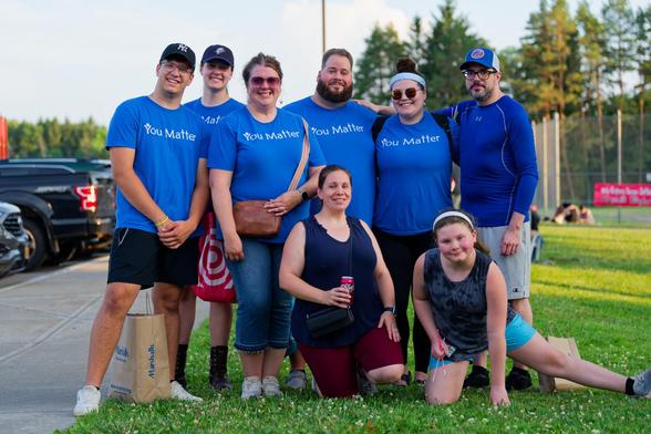 8 men and woman post together with a somewhat confused background, most of the people in back have blue shirts that say "You Matter"
