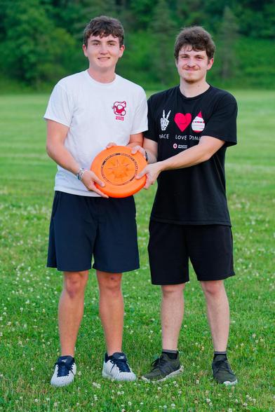 Two young men hold an orange frisbee; on the left with a white shirt that reads CORNELL 2025 and on the right with a black shirt advocating blood donations.  they are on a close-cropped lawn with lovely clover with a darker hedgerow behind