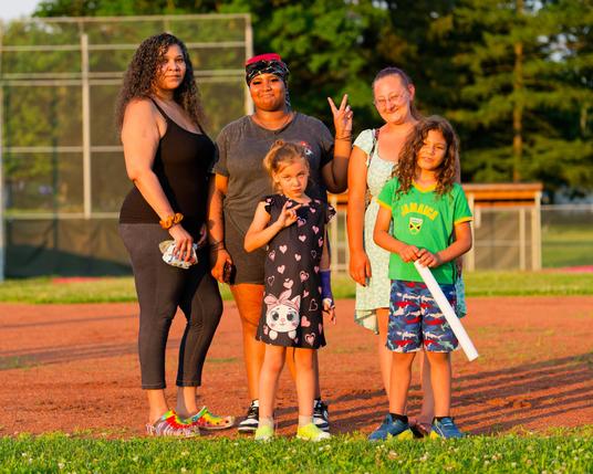 Three adult woman in the back and two girls in the front with a youth baseball field behind them