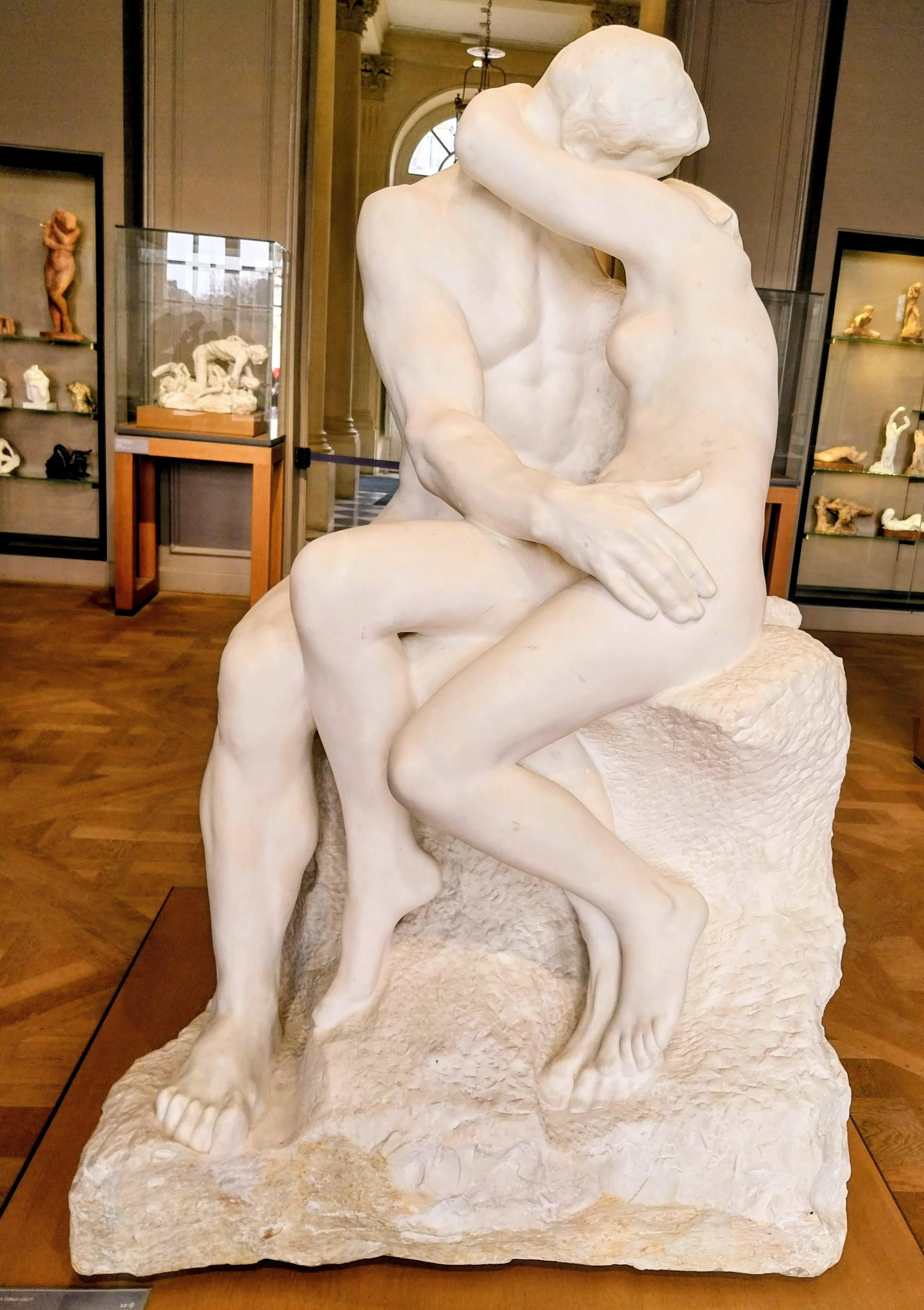 Skulptur eines eng umschlungenen Paares in weißem Marmor. Sie sitzen auf einem weißen Sockel und küssen sich. Im Hintergrund sind weitere Ausstellungsstücke in Vitrinen zu sehen.