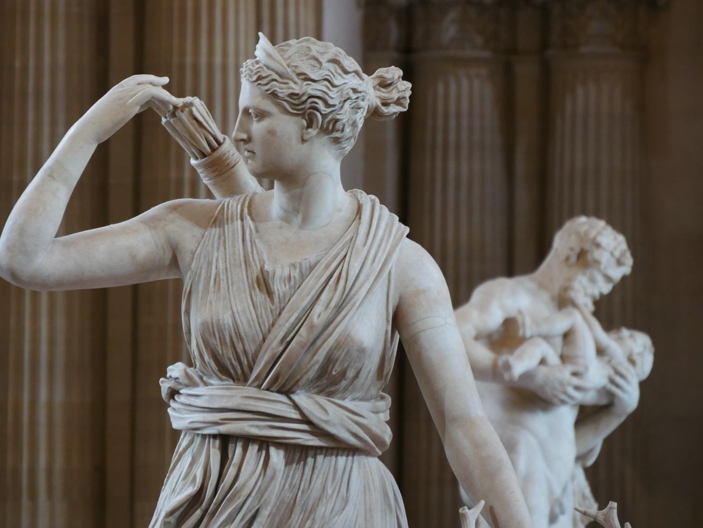 Photo aus der antiken Skulpturensmmlung des Louvre. Im Vordergrund ist die Jagdgöttin Artemis/Diana zu sehen, wie sie gerade dabei ist einen Pfeil aus dem Köcher zu ziehen. Artemis und die Hirschkuh. Bemerkenswert ist die feine Arbeit des Faltenwurfs ihres Kleids. Die Statue im Hintergrund zeigt den gealterten Satyr Silenos, den weisen Lehrer und Begleiter des Weingottes, der den jungen Dionysos liebevoll im Arm hält. Paris 2026