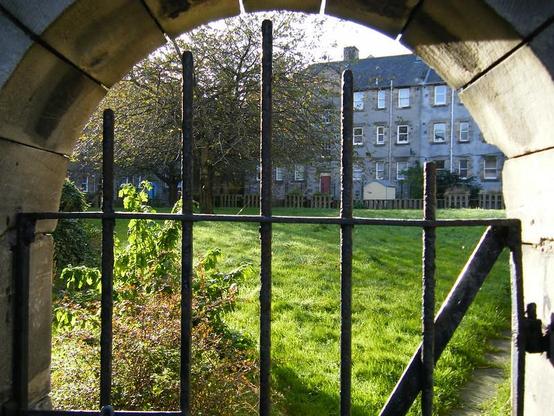 Looking through a stone archway, with a very old (and heavy!) iron gate. Beyond is a grassy courtyard and the rear of some older flats. Central Edinburgh, near the Pleasance.