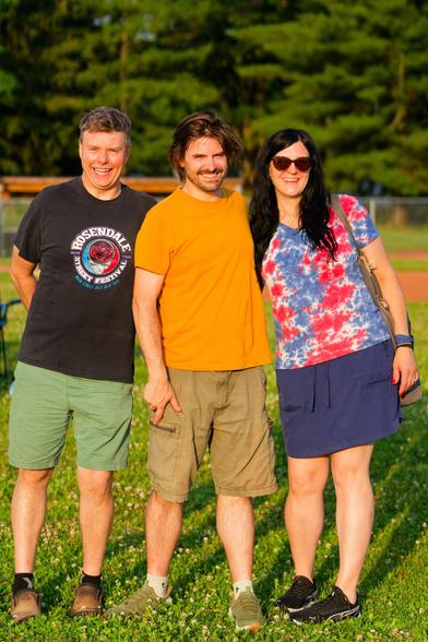 Three people on close cropped grass with evergreen trees in the background:  (left) man with green shorts and black shirt with circular logo and short straight hair (middle) man with solid yellow shirt and brown shorts, a beard and curly short and (right) woman with blue above-the-knees skirt, red and blue tie die shirt, dark hair well past shoulders and dark glasses