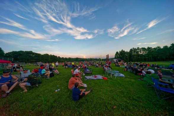 Lots of people are sitting in a large grass field under a blue sky with a few cirrus clouds with forest looming behind them and a red band of sky indicating imminent sunset