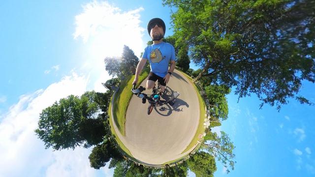 A photo from a 360 degree action cam of a silly man riding a bike that makes it look like he is riding on a tiny planet.