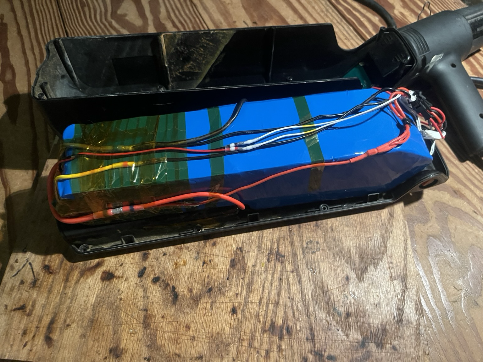 Battery pack wrapped in heat shrink and sitting in lower half of the case with upper half of the case laying beside. All the wires are connected using solder seal and a heat gun sits beside the pack.