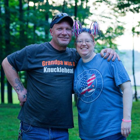 On the left a man with a dark shirt that reads "Grandpa was a Knucklehead" wearing a baseball cap and on the right a woamn with a light blue shirt with an American flag-based symbol inscribed in a double circle and a headdress with red, white and blue streams on a lawn with a row of trees and an open scene including a lake behind