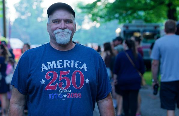 Man with baseball cap and neatly trimmed beard and mustache wears blue shirt that reads AMERICA 250 years 1976 - 2026 with a crowd of people behind him blurred out and hazy trees in the far background
