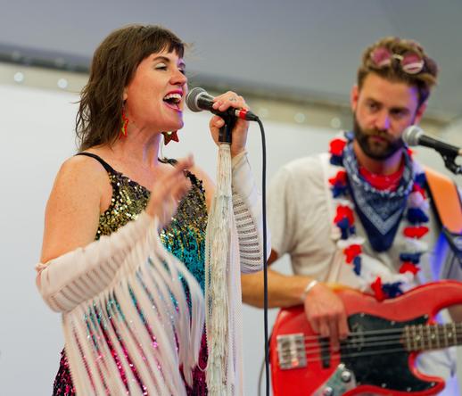 On the left a woman singer her hear out into a microphone with shoulder-length hair and a sparkly dress wearing things on her arm with numerous white strips trailing from them, and on ethe right a man playing a red guitar with a white shirt and a red white and blue leah and multiple scards, short hair and round glasses on his head
