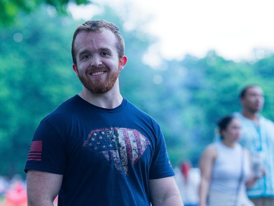 Smiling man with short hair and neat beard and mustache with a dark blue shirt with a diamond area with starts strips and a blue field with white flags like the American flag and behind him there is a couple walking blured out and a wall of trees in the haze