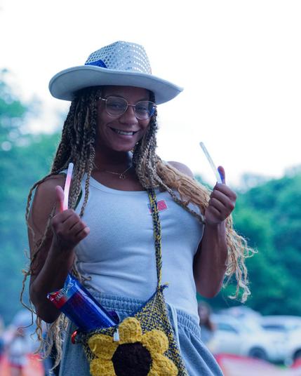 Black woman with white cowboy hat and shirt and long dreadlocks with a sunflower powers holding two light sticks with glasses and a big style:  behind her are blurred out cars near the bottom, hazy trees, and a blown out sky