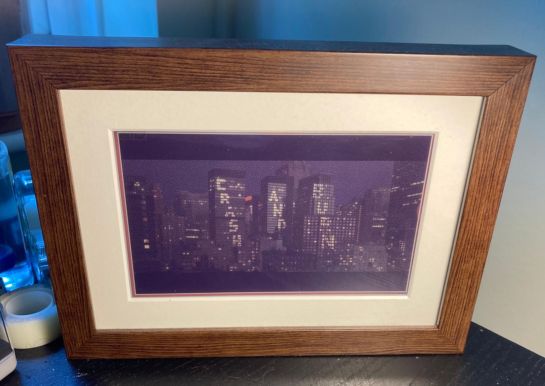 Photograph of a framed e-ink very slow movie player display, sitting on a black desk.  The frame is showing a frame from the film hackers, in this case the words "crash and burn" spelled out in the windows of skyscrapers in NYC.