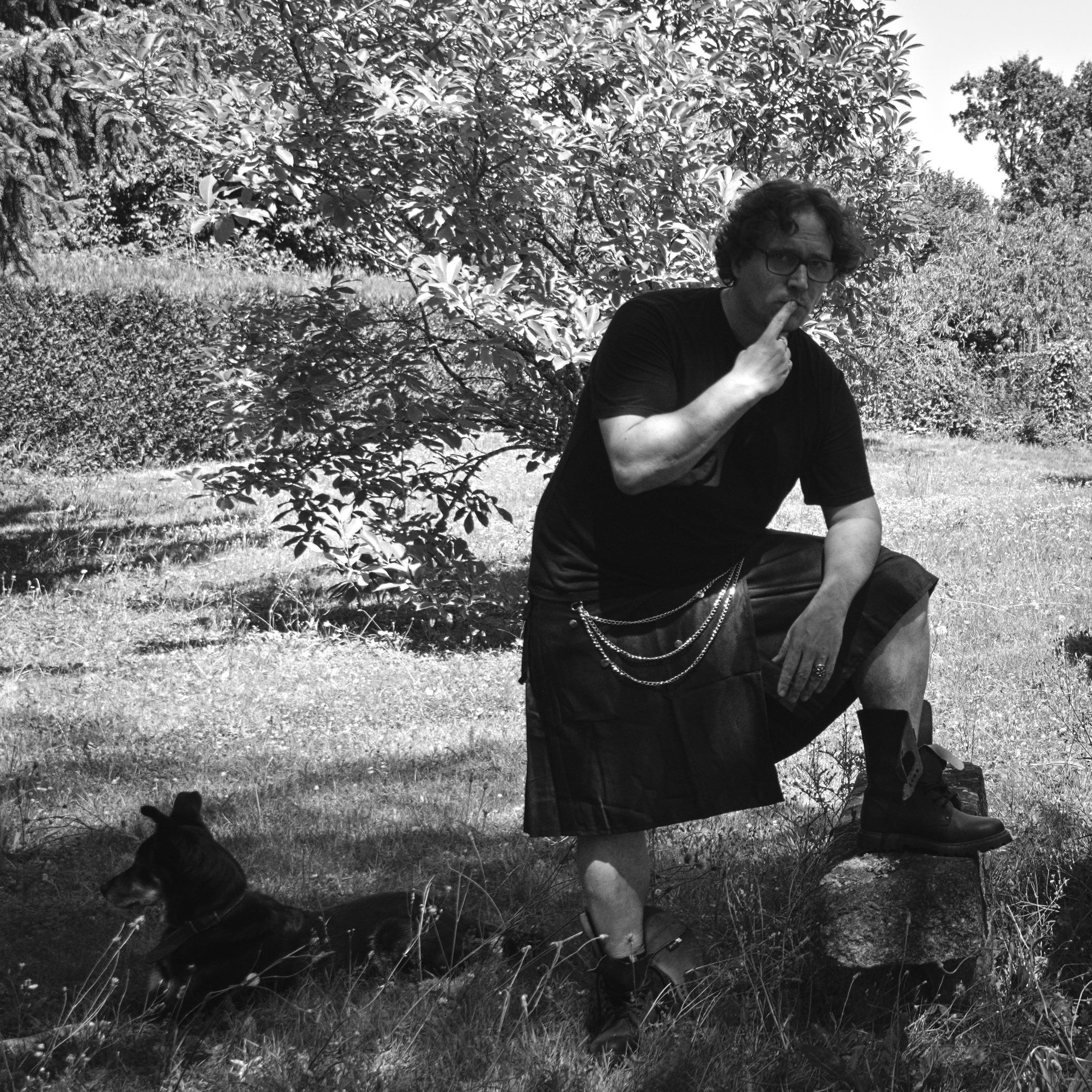Photo noir et blanc d'un chien allongé dans l'herbe au pied d'une personne portant un kilt avec trois jolies chaînes sur le devant, appuyant son coude sur sa jambe posée sur un petit banc de pierre, un doigt sur les lèvres dans une effroyable tentative de minauderie