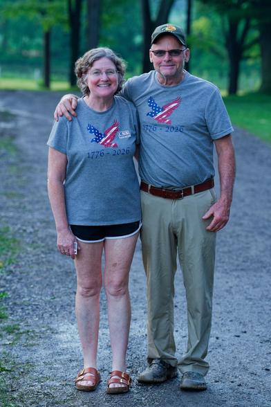 On the left a woman wearing shorts with short hair, on the right a main a head taller with a baseball cap and light colored pants and a brown belt holding the woman around her shoulder.  Both are wearing grey shirts with a picture of an eagle with stars and stripes.  They are standing on a path that curves to the left in the distance and has the stems of large trees in the background