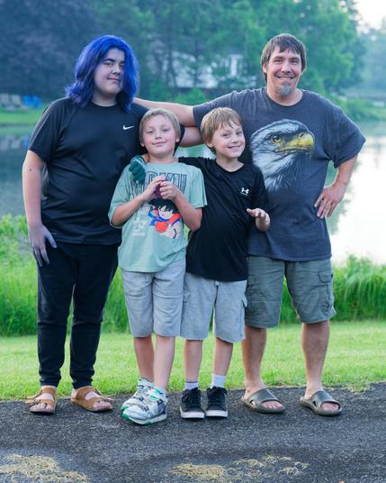 Left to right a person with blue hair in a black nike shirt and black pants with his arms locked with a guy on the far right who has short hair and a beard and a dark grey shirt with a eagle print and lighter shorts.  Between them two smiling children, one with an anime shirt and another with a dark shirt and both wearing light shirts.  They are standing on the edge of an asphault basketball court and behind them is a lawn and a pond with houses on the far end and water vegetation on the close …