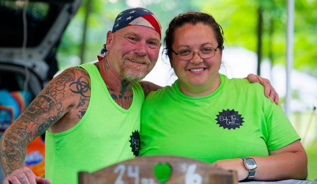 On the left a man wan wearing an American flag bandanna, tattooed arm and a light beard and mustache and on the right a woman who i think has her hair tied up,  both are wearing green shirts with the logo of their business.  Both the man and woman have beautiful smiles and the man has his arm around the shoulder of the woman.