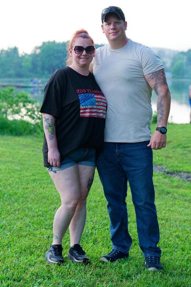 Man and woman stand on close cropped grass in front of large pond with trees on the far shore.  On the left the woman is wearing shorts and has a black shirt with an American flag and dark glasses and her hair tied up,  the man on the right is a head taller and has a white shirt with blue pants and  watch and a billed cap with glasses on top.  both have tattoos on their arms