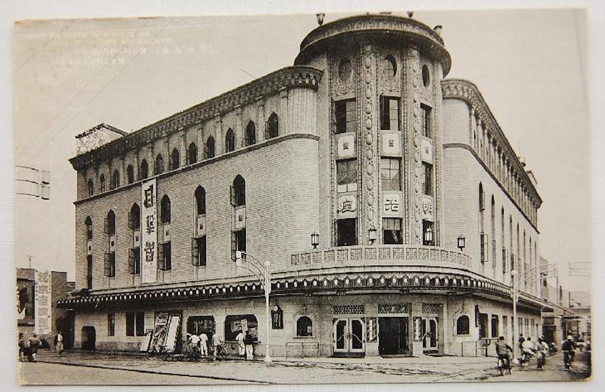 Architectural description: The Meiji-za Theater in Seoul

A monumental, multi-story corner building blending 1930s modernism with historicist elements. Its most prominent feature is a massive, semi-cylindrical corner tower that smoothly connects the two intersecting street facades.

Ground floor & entrance: The main entrance sits at the base of the rounded tower, sheltered by a heavy, dark marquee that extends across the facades, visually separating the ground floor from the upper levels.

The tower: Above the marquee, a curved balcony with a stone balustrade wraps around the first floor. The tower features high, narrow windows separated by vertical decorative relief strips. The top level displays small, circular (oeil-de-boeuf) windows, crowned by a flat, circular roof dome.

The wings: Branching from the tower, the facades feature a rhythmic window arrangement. The lower floors have tall, lancet (Gothic-style) windows with open dark shutters. The top floor is lined with a dense, continuous arcade of small, semi-circular windows right below the cornice.