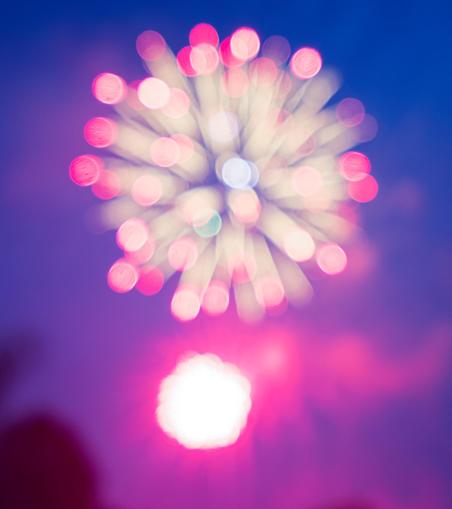Out of focus image of bright colored lights with white trailers radiating out from a point with a blown out ball of red light below and a red-lit clouds down low,  dark trees to the lower right corner and an overall dark blue background