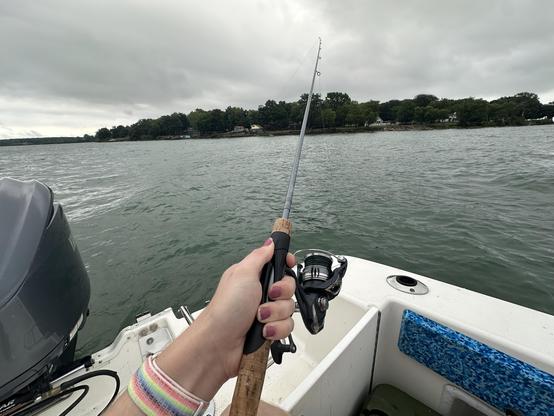 PoV of me fishing. I'm holding the spinning reel with my left hand with very worn smoky red nail polish. An outboard engine is to the left. The sky is cloudy and the water is calm off of Kelly's Island in Lake Erie