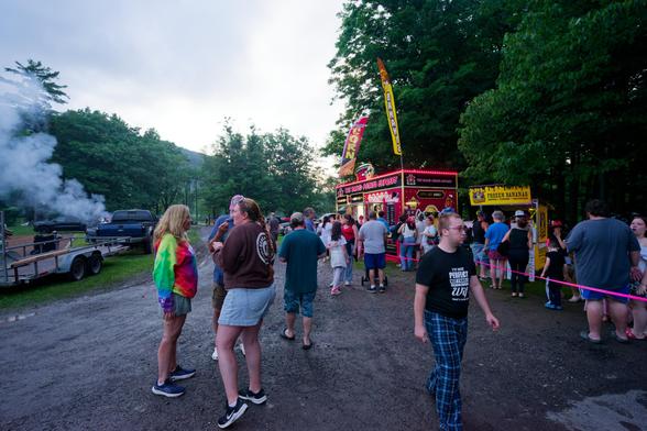 To the left a plume of smoke is rising behind a black pickup truck with a trailer, i have no idea if it is somebody cooking or from a wildcat firework;  in the middle a crowd fills a dirt trail, on the left people are queued behind a purple tape for a larged baked potato truck with banners and a small frozen banana booth with a row of wide and tall trees behind.