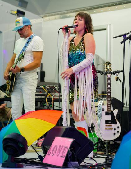 On the left a man with a white t-shirt and white pants and blue cap and electric guitar with a blue strap.  A little right of center find a woman with shoulder-length hair and sparkly multi-colored slinky dress and arm warmers with long white strands trailing below.  She sings into a microphone but their is a white electric guitar right next to her.  In front a rainbow umbrella, a fan, a monitor sapeak and a pink sign that reads LOVE.