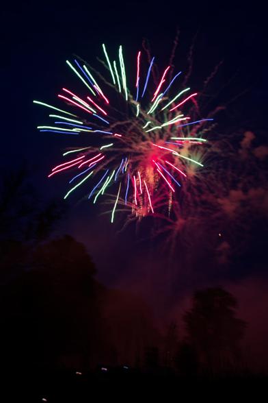 The upper half of the frame is mostly filled by red white and blue streaks radiating out from the center (but starting halfway out) under a dark blue sky:  behind and below that are streaks and puffs of smoke and under that the silhouettes of trees, a big cloud of smokes and a few motion streaked lights.
